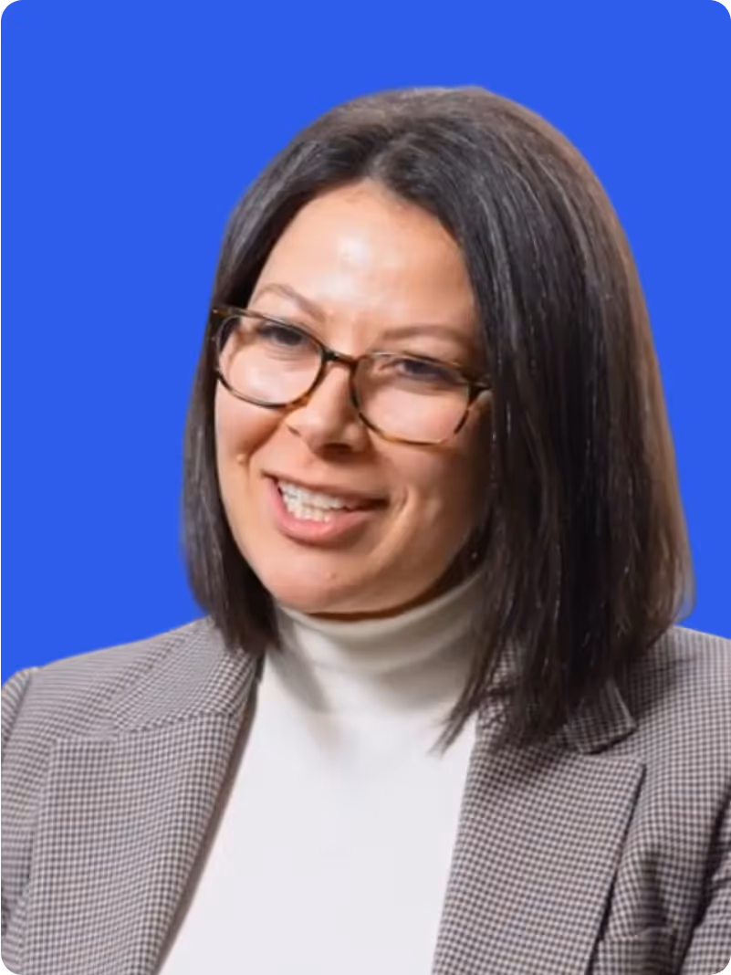 Woman with straight shoulder-length dark hair wearing glasses, a white turtleneck, and a checkered blazer smiling against a blue background.